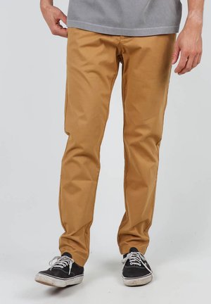 Chinos beige ajustés avec une texture lisse. Comprend des poches latérales et une ceinture sécurisée. Associés à des baskets noires et un t-shirt gris.
