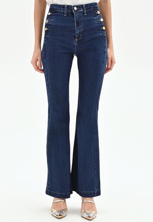 Jean large taille haute bleu foncé avec des boutons dorés sur les poches, porté avec des talons pointus argentés sur un fond blanc.