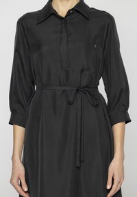 Czarna satynowa sukienka z kołnierzykiem, półguzikowym zapięciem, kieszenią na piersi i wiązanym paskiem w talii. Rękawy trzy czwarte.