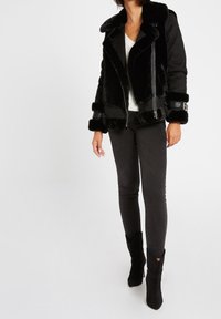 Černá kabát z umělé kožešiny s límečkem, zapínáním na zip a přezkami na rukávech. Nositý přes bílý top a černé džíny.