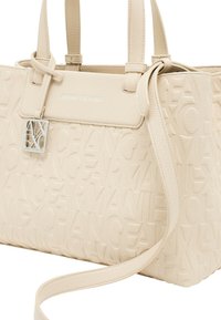 Sac à main beige avec motif de logo en relief, surface en cuir texturé, double anses et bandoulière amovible avec détail en métal sur le logo.
