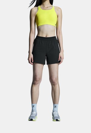 Kvinde står iført gul sports-bh, sorte atletikshorts, lyseblå sokker og hvide løbesko på hvid baggrund.