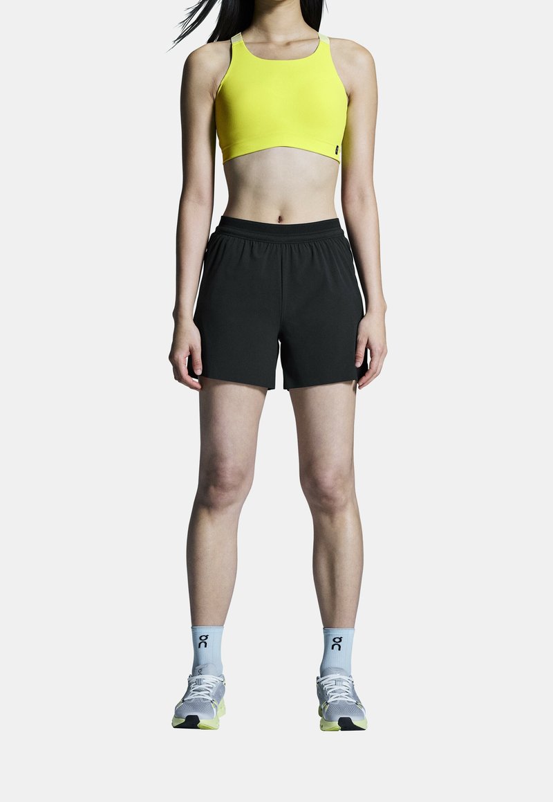 Femme debout portant un soutien-gorge de sport jaune, un short de sport noir, des chaussettes bleu clair et des chaussures de course blanches sur fond blanc.