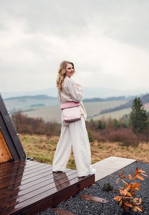 Hellrosa Umhängetasche mit flachem Design und verstellbarem Riemen, kombiniert mit lockeren weißen Hosen und einem hellfarbenen Oberteil, vor natürlichem Hintergrund.