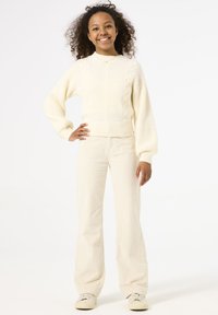 Suéter de punto crema con cuello alto y patrón de trenzas, combinado con pantalones de campana crema. La modelo lleva zapatillas ligeras y está de pie con las manos en las caderas.