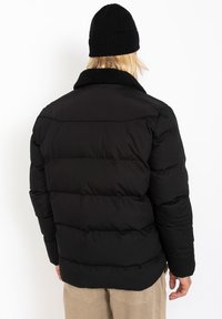Sort pufferjakke med faux fur-krave, der har et quiltet design og tykt polstring for varme. Bagfra fremvises en afslappet pasform og elastiske manchetter.