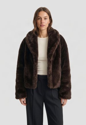 Femme portant un manteau en fausse fourrure marron foncé par-dessus un haut blanc et un pantalon noir, debout devant un fond clair uni.