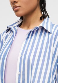 Νεαρός που φοράει μπλε και λευκό πουκάμισο με κάθετες ρίγες πάνω από ένα ανοιχτό μωβ μπλουζάκι με στρογγυλή λαιμόκοψη, με πλεξούδες στα μαλλιά.