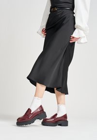 Mocassini in pelle bordeaux con una spessa suola in gomma nera, indossati con calzini bianchi scollopati e abbinati a una gonna in satin nera.