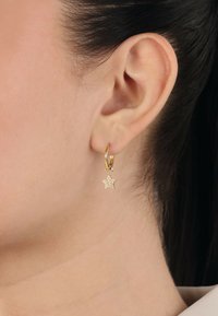Pendiente de aro dorado con un charm de estrella colgante adornado con rhinestones claros, textura suave y acabado pulido.