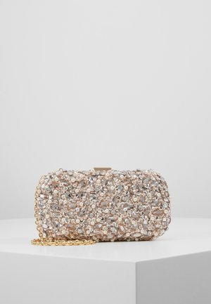 Pochette ornée de perles, cristaux transparents et argentés, avec une bandoulière chaîne dorée, présentée sur un socle blanc sur fond gris clair.
