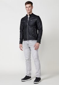 Chaqueta de cuero negra con cuello alzado, bolsillos con cremallera y detalles texturizados. Llevada sobre una camisa blanca y combinada con jeans grises claros y zapatillas negras.
