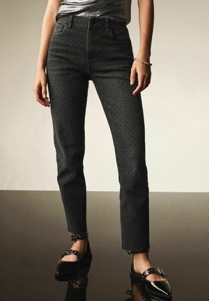 Jeans in denim nero con vita alta, decorati con piccole borchie, con una vestibilità slim e orli sfrangiati. Abbinati a ballerine nere con embellimenti.