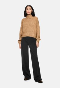 Brauner gestrickter Rollkragenpullover mit geripptem Saum, kombiniert mit schwarzen weiten Hosen. Das Model steht vor einem einfarbigen Hintergrund.