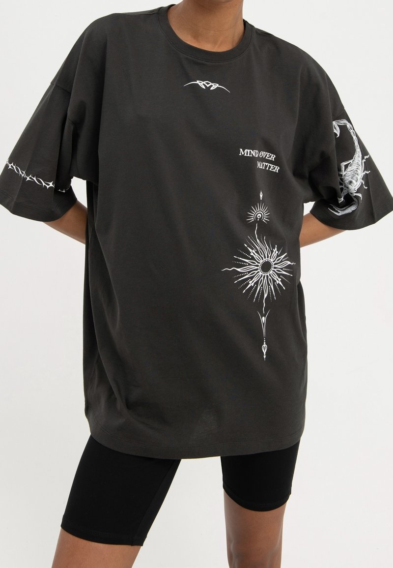 Personne portant un t-shirt noir ample avec des motifs blancs de soleil, scorpion, fil barbelé, et le texte "MIND OVER MATTER", assorti à un short de cyclisme noir.