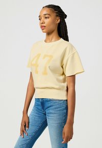 Felpa gialla a maniche corte con una grafica testurizzata "47" sul davanti, caratterizzata da un orlo a costine e un tessuto morbido. Abbinata a jeans blu.
