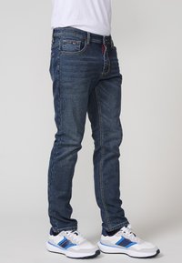 Vaqueros de mezclilla en azul oscuro con un corte slim. Las características incluyen cinco bolsillos, costuras en contraste y un detalle de etiqueta roja. Combinados con zapatillas blancas.