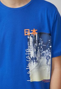 Koninklijk blauwe katoenen t-shirt met een grafische print van de lichten van Tokio, met oranje tekst en een contrasterend monochroom beeld.