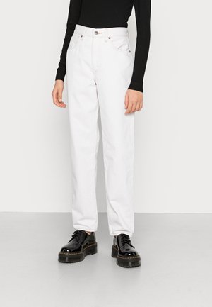 Osoba ubrana w białe dżinsy z wysokim stanem, czarną bluzkę z długim rękawem oraz błyszczące czarne buty na platformie, stojąca na jednolitej podłodze.