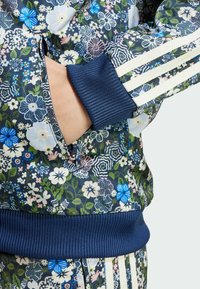 Blommig sportjacka med marinblå ribbade muddar. Har en fodrad ficka med dragkedja och livliga färger inklusive blå, rosa och gröna blommönster.