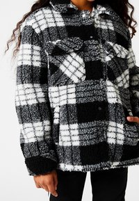 Chaqueta de forro polar a cuadros en blanco y negro, con cierre de botones en la parte delantera, dos bolsillos en el pecho y material texturizado y suave.