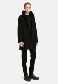 Zwarte jas van gestructureerde stof met een capuchon, twee knopen en zijzakken. Gecombineerd met zwarte slim-fit broek en stevige zwarte laarzen.