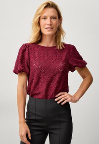 Bordeaux blonde top med puffærmer, der har et blomstermønster og en tekstureret overflade, parret med sorte tætsiddende bukser.