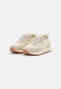 Pár béžových a krémových sneakers s gumovými podrážkami, semišovými akcenty, vzorovanými tkaničkami a brandingem po straně, vystavených na bílém pozadí.