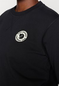 Felpa nera in cotone, con collo rotondo, cuciture piatte e una toppa circolare con un logo di volpe e testo.