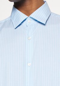 Camicia azzurro chiaro con sottili righe verticali bianche, colletto a punta e tre bottoni bianchi visibili sul davanti.