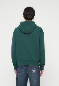 Felpa verde con cappuccio, dal taglio rilassato, in tessuto testurizzato e con tasca a marsupio, indossata con jeans in denim blu con una toppa a forma di stella.