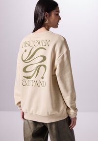 Sudadera beige con un gran diseño gráfico verde y el texto "DESCUBRE Y EXPANDE" impreso en la parte de atrás. Tela suave, corte relajado.