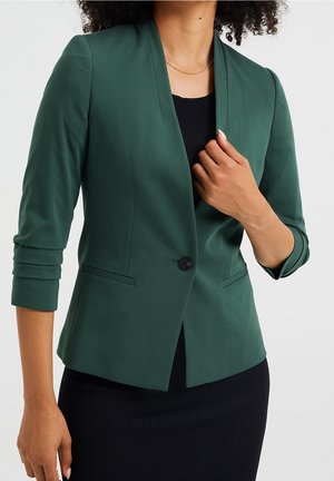 Femme portant un blazer vert foncé cintré à manches trois-quarts sur une robe noire côtelée, main ajustant le devant du blazer.