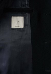 Manteau noir ouvert pour montrer la doublure sombre avec une étiquette de marque blanche indiquant "MNG Man" et les détails de taille cousus à l'intérieur.