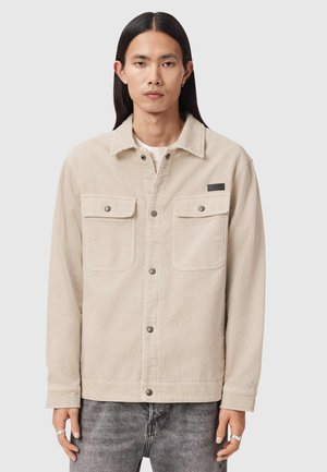 Mężczyzna o długich czarnych włosach w beżowej kurtce sztruksowej z zapięciami na zatrzaski i kieszeniami na piersi, w połączeniu z szarymi, spranymi dżinsami.