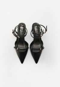 Talons aiguilles en cuir vegan noir avec des brides de cheville et des accents en métal doré. Texture lisse et design minimal.