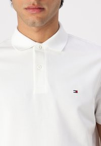 Balanšinės medvilnės balta polo marškinėliai su klasikiniais apykaklėmis, ant kairio krūtinės yra mažas raudonas ir mėlynas siuvinėtas logotipas.