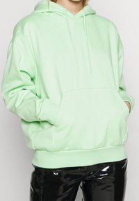 Sudadera con capucha verde menta, con un bolsillo canguro, cordones ajustables y puños acanalados, combinada con pantalones negros brillantes.