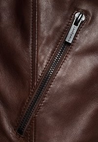 Brązowa skórzana kurtka z widocznym czarnym zamkiem błyskawicznym i szwami. Skóra ma gładką teksturę z subtelnymi detalami ziarna.