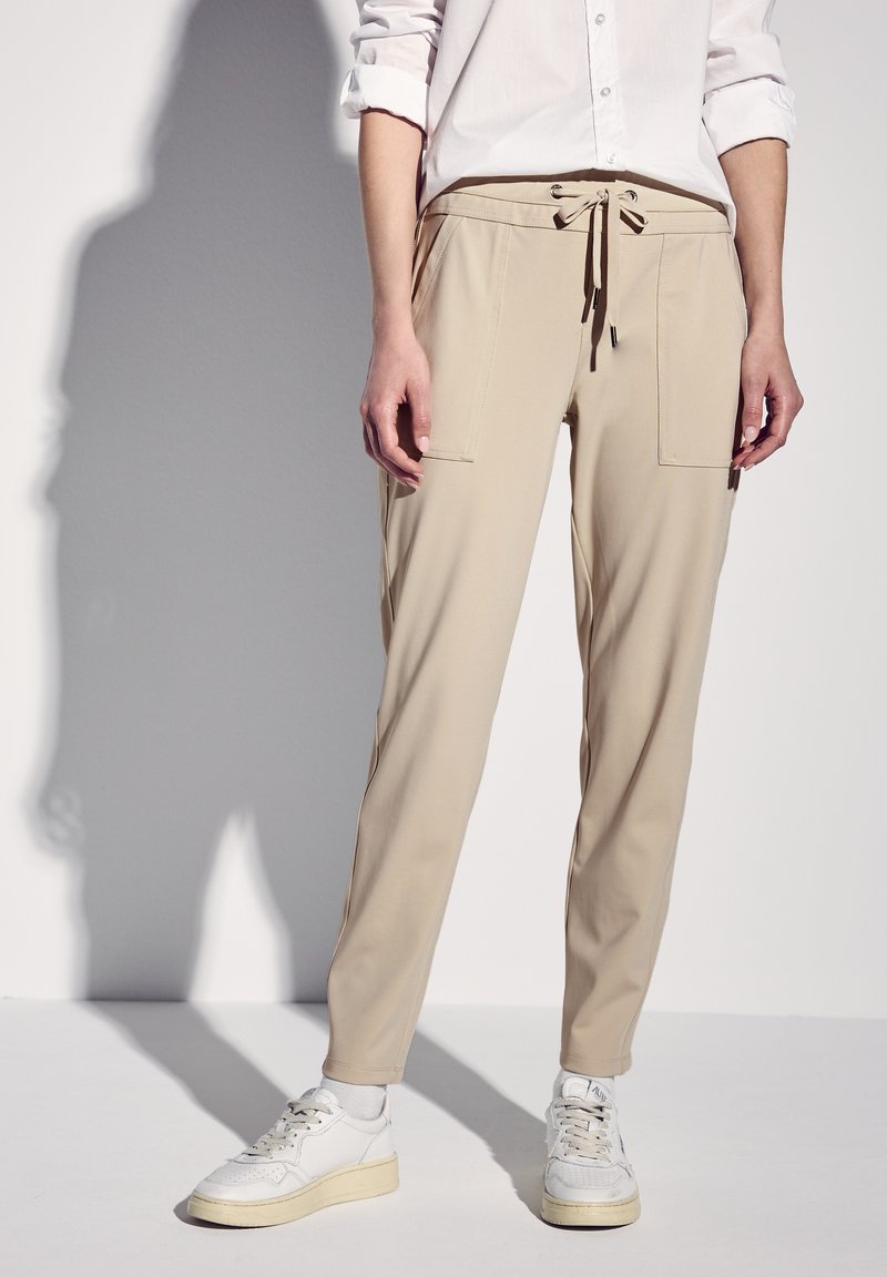 Person, die beige Kordelzughosen mit Vorderentaschen, ein weißes, hochgekrempeltes Hemd, weiße Socken und weiße Turnschuhe trägt und vor einem hellen Hintergrund steht.