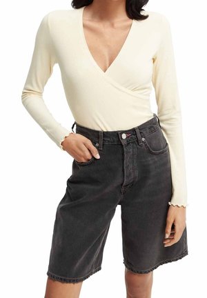 Vrouw draagt een crèmekleurige, lange mouw V-hals top, ingestopt in een high-waisted, losse zwarte denim broek tot aan de knieën.