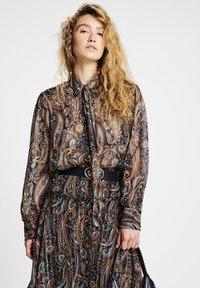 Long-sleeve, patterned blouse with a tied neck, featuring intricate paisley designs in browns, blues, and blacks. Accompanied by a black belt.