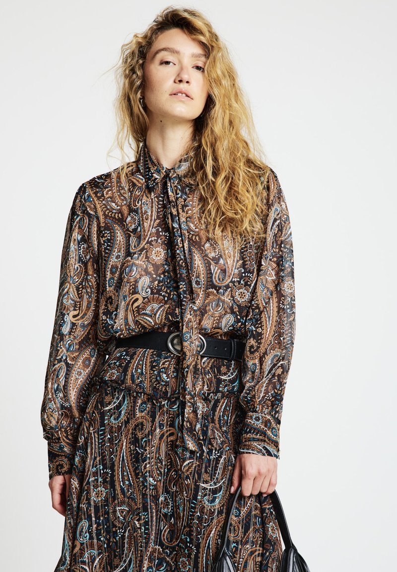 Long-sleeve, patterned blouse with a tied neck, featuring intricate paisley designs in browns, blues, and blacks. Accompanied by a black belt.