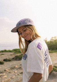 T-shirt blanc avec graphique floral bleu et étiquette violette ; casquette à motif violet clair. L'arrière-plan présente de la verdure et un paysage flou.