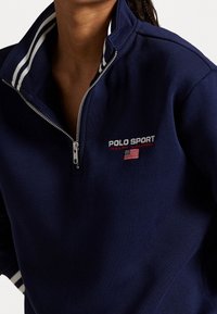 Navy blue half-zip sweatshirt featuring white stripes on the collar, embroidered "POLO SPORT" logo, and a silver zipper detail.