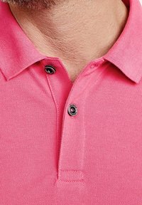 Różowa koszulka polo o miękkiej fakturze, krótki kołnierzyk i zapięcie na dwa guziki z wyraźnymi czarnymi akcentami guzika.