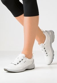 Bílé sportovní boty s pletivovým svrškem, stříbrnými akcenty a kulatou špičkou. Mají polstrovanou podrážku a design na šněrování.
