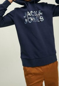 Felpa blu navy con logo frontale "JACK & JONES" stampato in multicolor. Presenta un cappuccio con cordino e un orlo a costine per una vestibilità aderente.