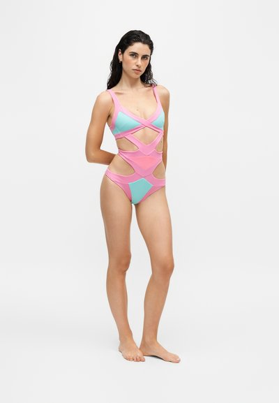 Woman standing barefoot wearing a pink and teal cut-out one-piece swimsuit on a plain white background.