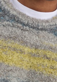 Pull en peluche avec une surface texturée en gris, présentant des rayures horizontales jaunes et bleues. Encolure côtes avec une couche intérieure blanche.